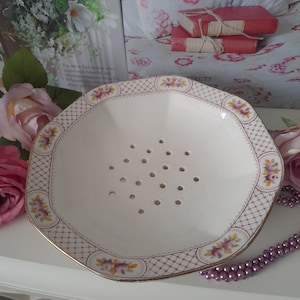 May include: A white ceramic strainer with a scalloped edge and a floral design. The strainer has a grid of small holes in the bottom and is decorated with purple and yellow flowers.
