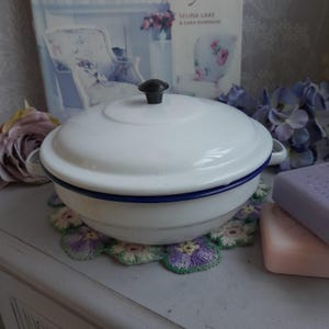 Może przedstawiać: Białe naczynie do zapiekania z emalii z czarnym pokrętłem na pokrywce i niebieskim obramowaniem. Naczynie stoi na kwiatowej podkładce. Książka i kwiaty są w tle. Naczynie ma średnicę około 20 cm.