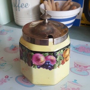 Pode incluir: Um açucareiro hexagonal de cerâmica amarelo com tampa de prata e um desenho floral com uvas, maçãs e folhas. O açucareiro está sobre uma mesa com um padrão floral.