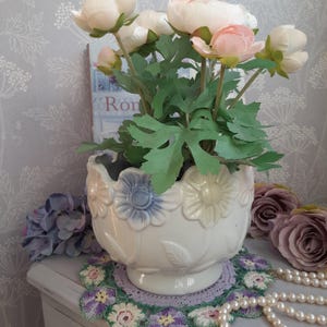 Pode incluir: Um vaso de flores de cerâmica branca com borda ondulada e detalhes de flores em relevo em azul e amarelo contém flores artificiais rosa e brancas com folhas verdes. Uma toalha de mesa de crochê e pérolas de imitação estão em primeiro plano.