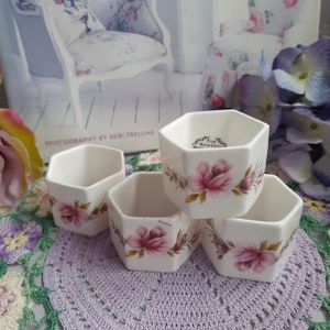 May include: Set of four white hexagonal ceramic napkin rings with a floral design featuring pink roses and green leaves. The rings are marked "Fine Bone China Made in China."