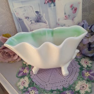 Puede incluir: Un plato de cerámica blanco con borde festoneado y tres patas pequeñas. El borde interior es de color verde claro. El plato está sobre un tapete morado con bordados florales.