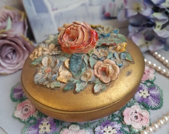 Puede incluir: Un joyero redondo de tono dorado con una tapa decorativa adornada con un grupo esculpido de flores y hojas multicolores en tonos rojo, rosa, azul y verde. La base del recipiente tiene un acabado metálico dorado liso y uniforme.