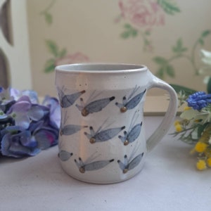 Puede incluir: Una taza de cerámica blanca con un diseño azul y dorado. La taza tiene un asa y está decorada con un patrón de pájaros estilizados.