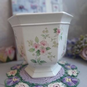 May include: White ceramic planter with a hexagonal shape and a floral design featuring pink and green flowers. The planter has a wide base and a narrow top.