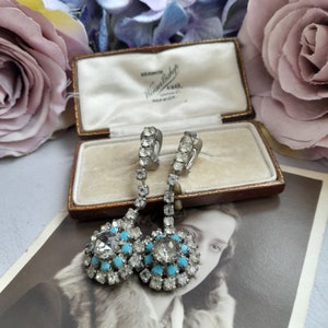May include: A pair of vintage dangle earrings with a silver tone finish. The earrings feature a cluster of clear rhinestones with a turquoise accent. The earrings are in a jewelry box with the text "Goldsmith Winifred Bishop London 4443 Norwich".