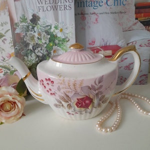 Può includere: Una teiera vintage con un design floreale rosa e bianco. La teiera ha un coperchio rosa e un bordo dorato. La teiera è appoggiata su una superficie bianca con una collana di perle.