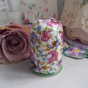 May include: A white ceramic jar with a floral pattern in shades of pink, purple, and yellow. The jar has a green rim and a white base.