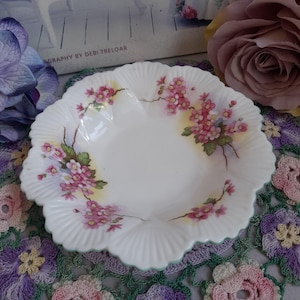 Może przedstawiać: Biały talerz z falistym brzegiem i kwiatowym wzorem. Talerz ma różowe kwiaty z zielonymi liśćmi i żółtymi akcentami. Obrzeże ma subtelne zielone wykończenie. Talerz jest prawdopodobnie porcelanowy lub ceramiczny.