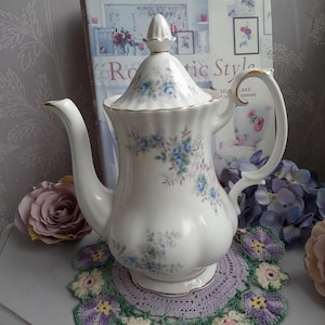 May include: A white porcelain teapot with a floral pattern of blue and purple flowers. The teapot has a gold trim and a handle. It is sitting on a crocheted doily.