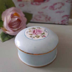 May include: A small, light blue ceramic trinket box with a gold trim. The lid features a painted design of pink roses and green leaves.