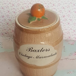 Può includere: Un barattolo di ceramica a forma di barile con una glassa marrone e un'etichetta bianca che recita "Baxter's Vintage Marmalade". Il coperchio è decorato con una foglia verde e un frutto arancione.