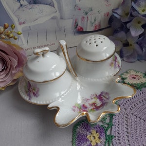 May include: A white porcelain salt and pepper shaker set with a floral design. The shakers are on a leaf-shaped tray with a gold trim.