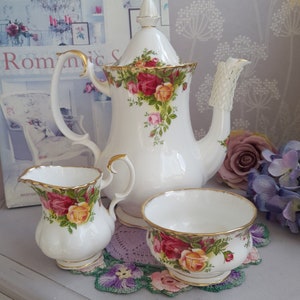May include: A white tea set with a floral pattern. The set includes a teapot, creamer and sugar bowl. The teapot has a lid and a handle. The creamer and sugar bowl have handles. The tea set is on a white doily.