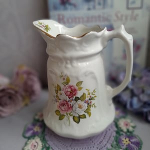 May include: A white ceramic jug with a floral design featuring pink and white roses, green leaves, and small purple flowers. The jug has a gold rim and a handle.