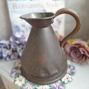 May include: A vintage copper jug with a flared spout and a curved handle. The jug has a dark patina and is sitting on a crocheted doily.