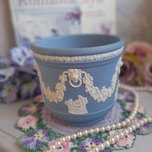 Puede incluir: Una maceta de cerámica azul claro con detalles en relieve blancos. La maceta presenta un borde decorativo, asas con forma de cabeza de león y figuras clásicas. Está sobre un tapete de ganchillo con un collar de perlas.