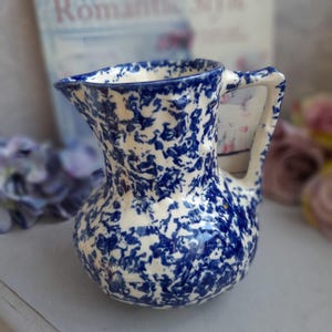 May include: A small ceramic jug with a white base and a blue splatter pattern. The jug has a handle and a spout. The design is reminiscent of vintage kitchenware. The jug is likely used for serving liquids.