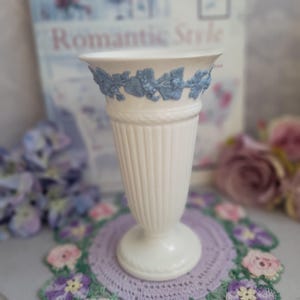 May include: A white ceramic vase with a fluted body and a flared rim. The rim features a blue grape and leaf design. The vase has a round base and a rope-like detail. A vintage home decor item.