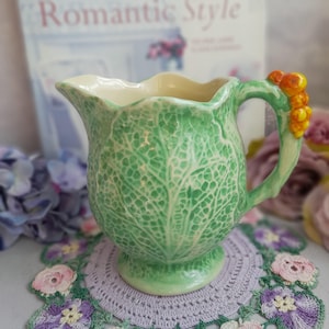 Peut inclure: Une cruche en céramique verte avec un motif de feuille de chou. La cruche a un intérieur crème et une anse ornée de baies orange. Elle repose sur un napperon violet avec des motifs floraux. L'arrière-plan comprend un magazine intitulé "Romantic Style."