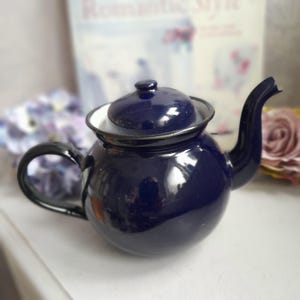 Op de afbeelding: Een donkerblauwe emaille theepot met een witte rand en een zwarte handgreep. De theepot heeft een rond lichaam en een tuit aan de zijkant.