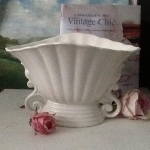 May include: A white ceramic vase with a fluted design and a scalloped rim. The vase has a decorative base with a scroll design. The vase is sitting on a white surface with two pink roses.