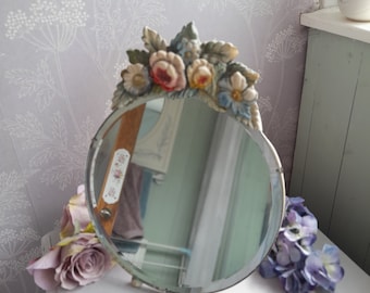 May include: A round vanity mirror with a floral design. The mirror has a decorative frame adorned with sculpted flowers in shades of pink, blue, and yellow. The mirror is supported by small feet and reflects the surrounding environment.