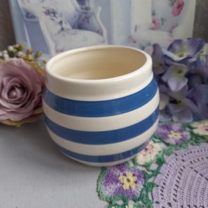 Può includere: Un barattolo di ceramica bianca con strisce blu. Il barattolo è rotondo e ha un'ampia apertura. È appoggiato su una superficie bianca con un pizzo all'uncinetto.