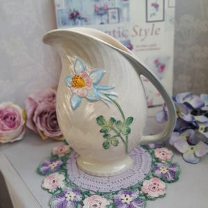 Può includere: Una brocca in ceramica color crema caratterizzata da un motivo floreale in rilievo dipinto a mano raffigurante un'aquilegia con petali blu, centro rosa e foglie verdi. La brocca presenta una finitura smaltata lucida e crepata, un manico curvo.
