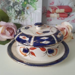 Può includere: Un porta burro vintage in porcellana con coperchio. Il piatto è decorato con un motivo floreale blu e oro su sfondo bianco. Il coperchio ha un pomo al centro.