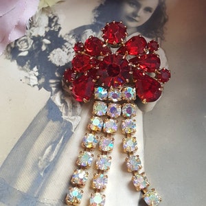 Peut inclure: Une broche vintage avec un groupe de strass rouges en forme de nœud, avec une cascade de strass clairs irisés qui pend.