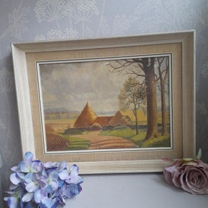 May include: A framed painting depicting a rural landscape with a thatched-roof farmhouse, trees, and a field. The painting is in a light wood frame with a cream-coloured mat.