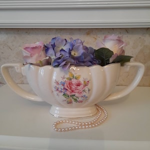 May include: A white ceramic creamer with a floral design and two handles. The creamer is filled with purple and pink flowers and a string of pearls is in front of it.