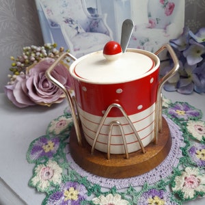 May include: A red and white polka dot sugar bowl with a silver lid and handle. The bowl is sitting on a wooden base with a crocheted doily.