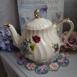 May include: A white teapot with gold trim and a floral pattern. The teapot has a lid with a gold knob and is sitting on a crocheted doily.