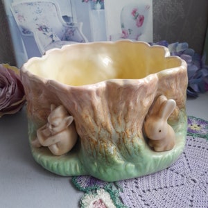 May include: A ceramic planter shaped like a tree trunk with a rabbit and a cat figurine on either side. The planter is glazed in shades of green, brown, and yellow.