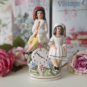 May include: A ceramic figurine of a boy and girl sitting on a clock. The boy is wearing a red hat and holding a flute. The girl is wearing a white dress and has a flower in her hair. The clock face is white with black numbers and hands.