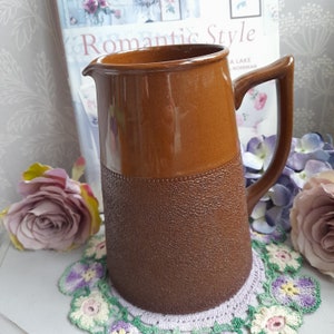 May include: A brown ceramic jug with a textured base and a smooth top. The jug has a handle and is sitting on a white crocheted doily.