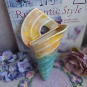 May include: A ceramic wall pocket in the shape of a seashell. The pocket is painted with a gradient of colors, transitioning from yellow and orange at the top to turquoise at the bottom. The opening is a curved shape, and the overall design is reminiscent of coastal decor.