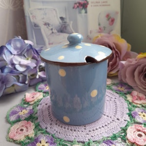 May include: A blue ceramic sugar bowl with a white polka dot pattern and a lid with a small spout. The bowl is sitting on a crocheted doily.