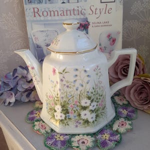 Puede incluir: Tetera de porcelana blanca con un patrón floral de flores rosas, azules y blancas. La tetera tiene un borde dorado alrededor de la tapa y el pico. La tetera está sentada sobre un encaje de ganchillo.