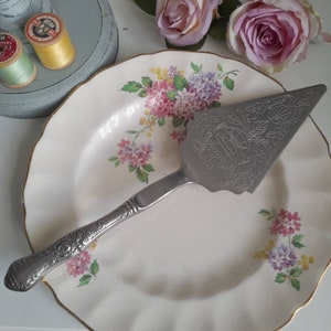 Puede incluir: Un servidor de pastel de plata con un diseño adornado en un plato blanco con un patrón floral. El plato tiene un borde dorado.