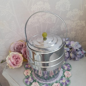 May include: A cylindrical chrome ice bucket featuring a tiered design, a polished metallic finish, a sturdy arched handle, and a small, lime green knob on the centre of the lid. The bucket has a smooth, reflective surface that captures light.