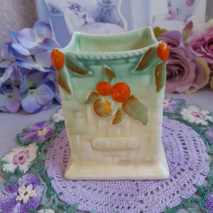 May include: A small, square, ceramic planter with a green and yellow glaze. The planter has a raised design of a woven basket with orange fruit and green leaves. The planter has the word "Spice" written on the front.