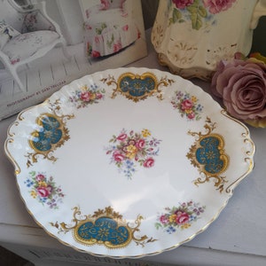 Puede incluir: Un plato para pastel blanco con borde festoneado y ribete dorado. El plato presenta un diseño floral azul y dorado con flores rosas, amarillas y blancas.