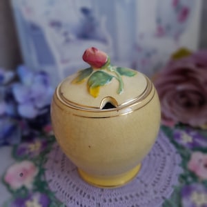 May include: A yellow ceramic sugar bowl with a lid decorated with a pink rose bud and green leaves. The bowl has a gold rim and base. The design is vintage and decorative.