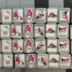 May include: A collection of rectangular, glittery, grey decorative tiles featuring various cartoon characters. The tiles showcase Hello Kitty, My Melody, and other characters with colourful designs and text. The tiles are arranged in a grid pattern.