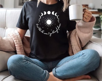May include: Black t-shirt featuring a white graphic of the sun surrounded by moon phases and the text "Just like the moon I go through phases." The shirt is worn with blue jeans and a light brown cardigan.