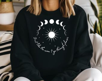 May include: Black crew neck sweatshirt with a white celestial design. The design includes a sun, moon phases, and the text "Just like the moon I go through phases". The text is in a cursive font. The sweatshirt is a casual style.