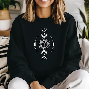 May include: Black sweatshirt with a white celestial design. The design features a sun with a face, surrounded by crescent moons and dotted lines, forming a symmetrical pattern. The sweatshirt has a crew neck.